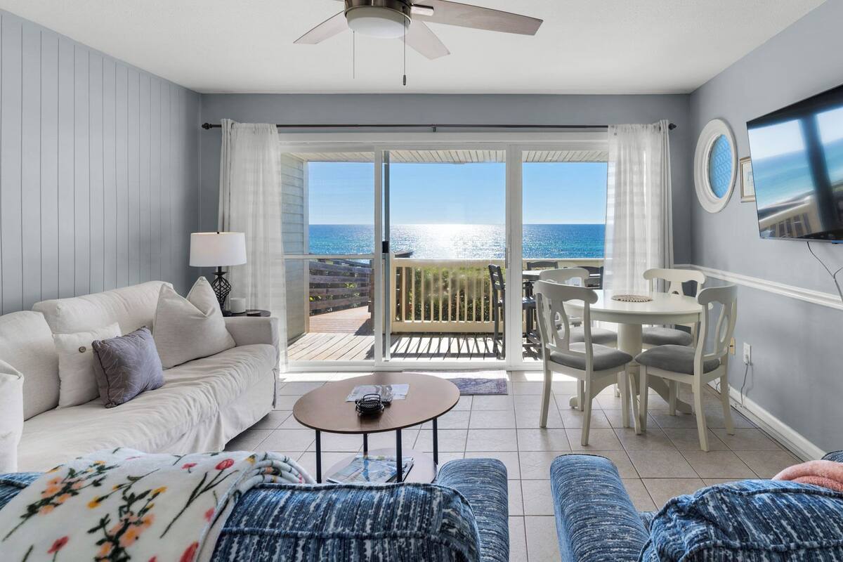 A living room with a sofa, coffee table, and ottoman, a dining area with chairs and a table, a TV mounted on the wall, and a balcony with a view of the sea.