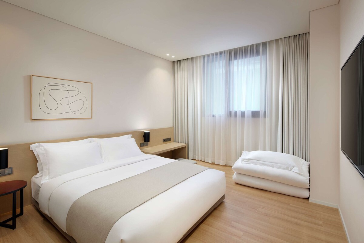 Een moderne hotelkamer met een groot bed, een nachtkastje, een flatscreen-tv en een raam met dunne gordijnen.