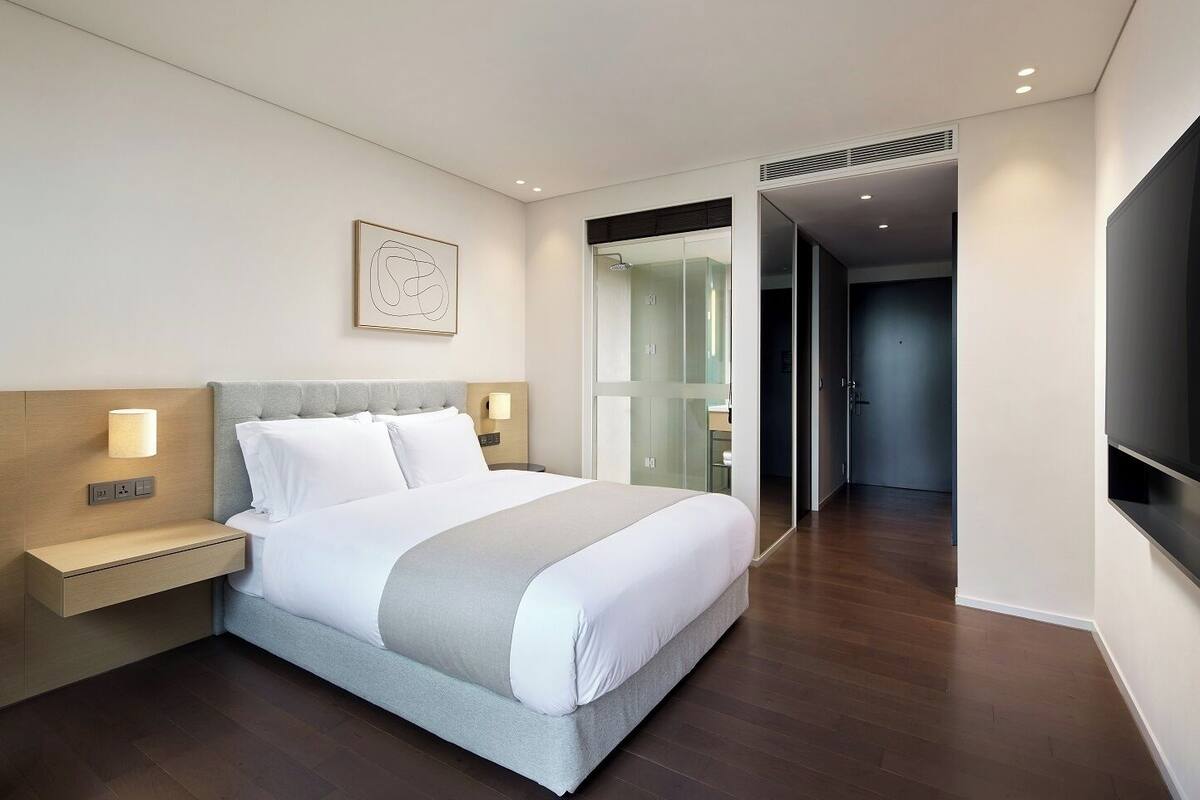 Quarto de hotel moderno com uma cama grande, mesas de cabeceira, televisão de tela plana e porta de vidro para um banheiro.