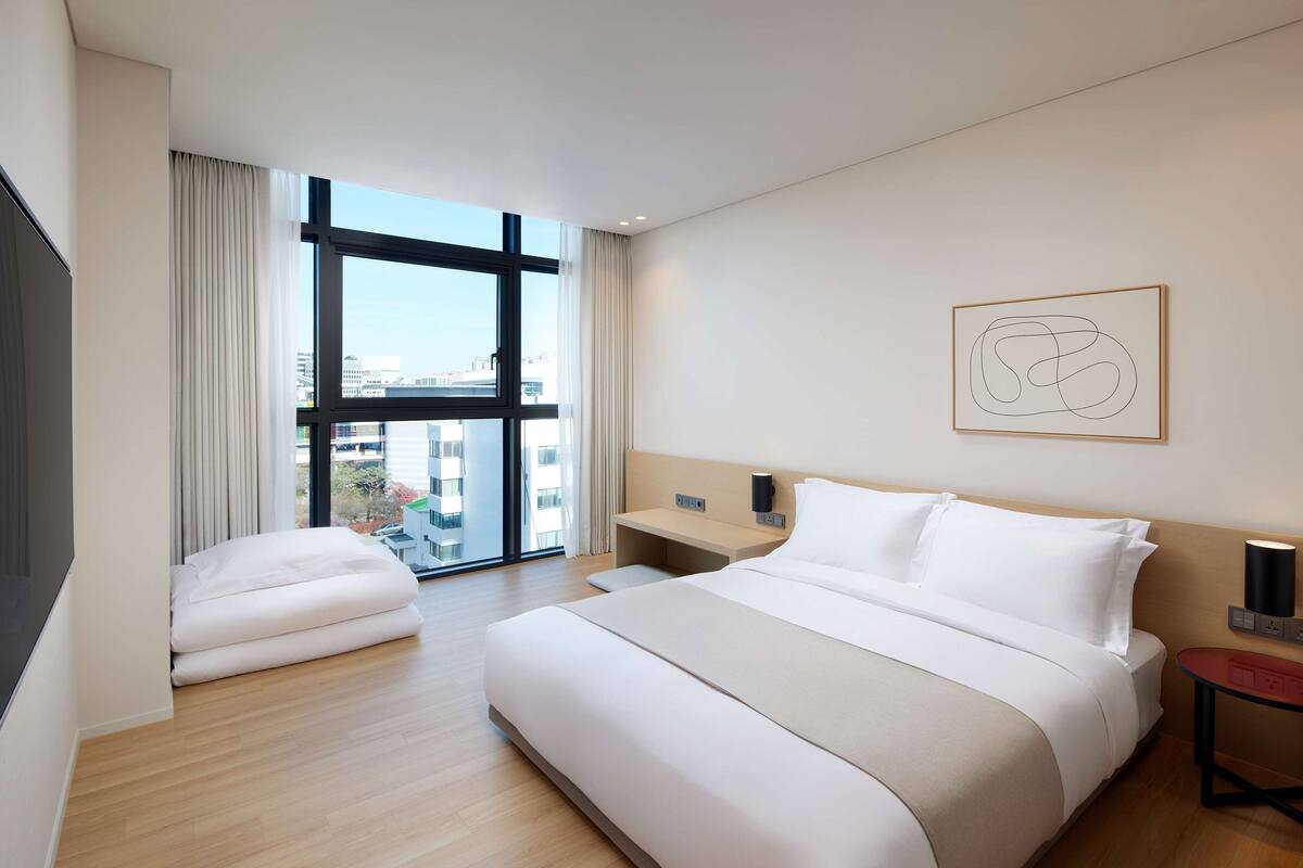 Quarto de hotel moderno com uma cama grande, escrivaninha e vista da cidade através de janelas do chão ao teto.