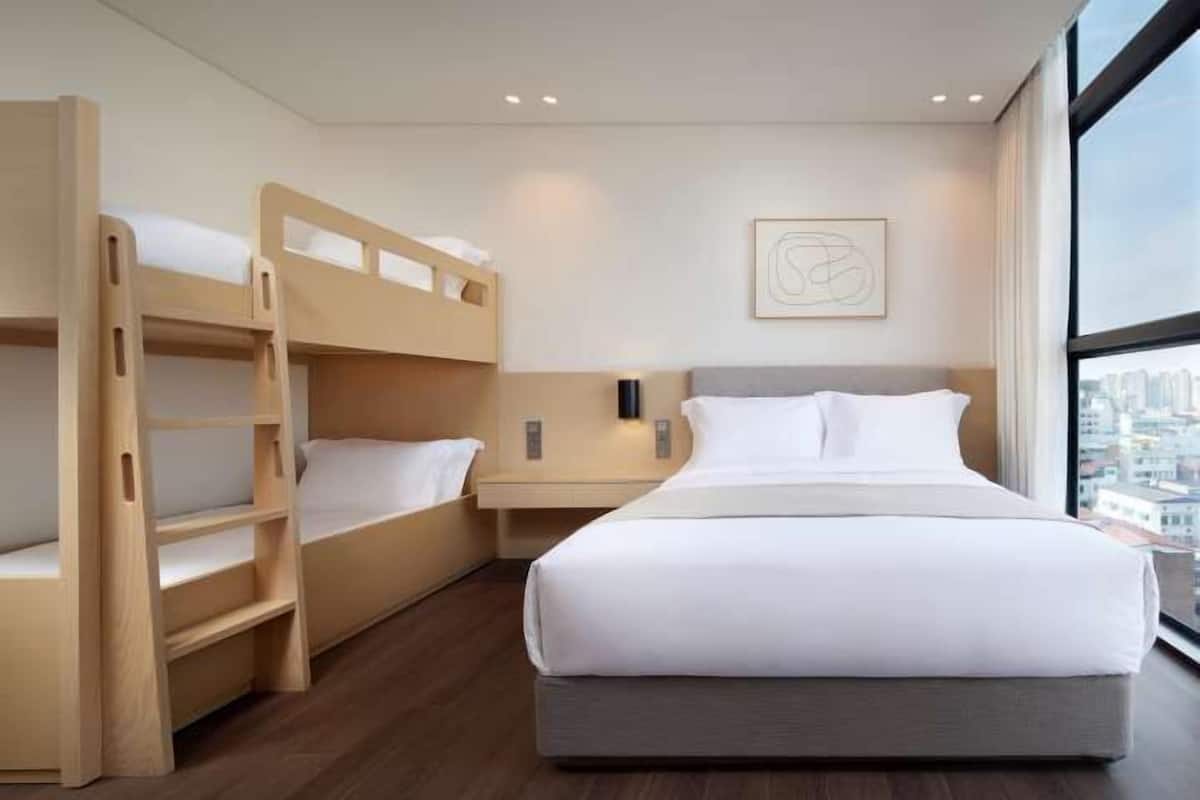 Quarto de hotel com uma cama grande, uma cama beliche menor, uma mesinha de cabeceira e vista para a cidade através de uma janela ampla.