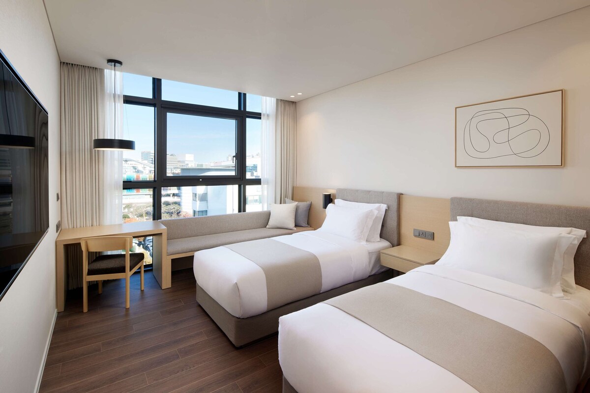 Een moderne hotelkamer met twee bedden, een bureau en een groot raam met uitzicht op de stad.