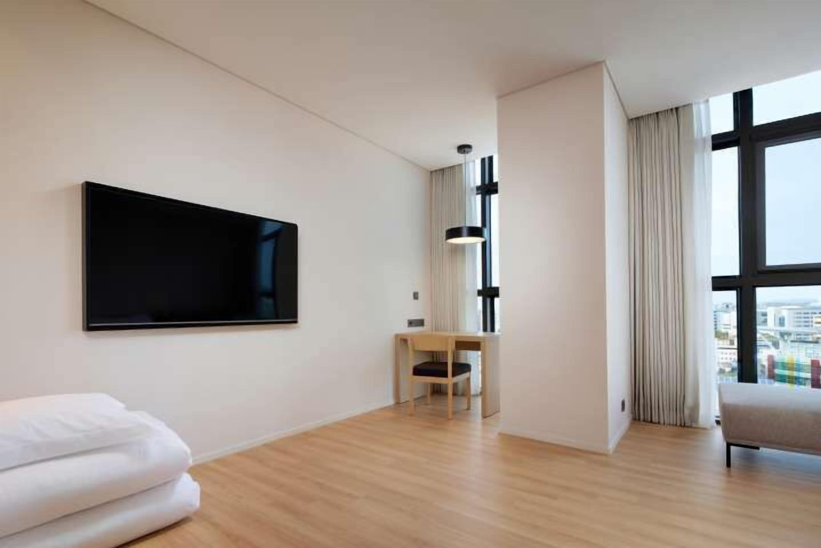 A modern hotel room with a large TV, a wooden floor, a small desk, and a view through large windows.