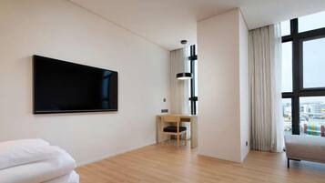 A modern hotel room with a large TV, a wooden floor, a small desk, and a view through large windows.