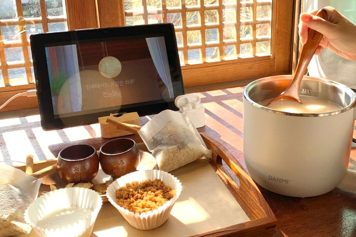A person stirring a liquid in a white mug on a wooden table with a laptop, a smartphone, and some snacks nearby.