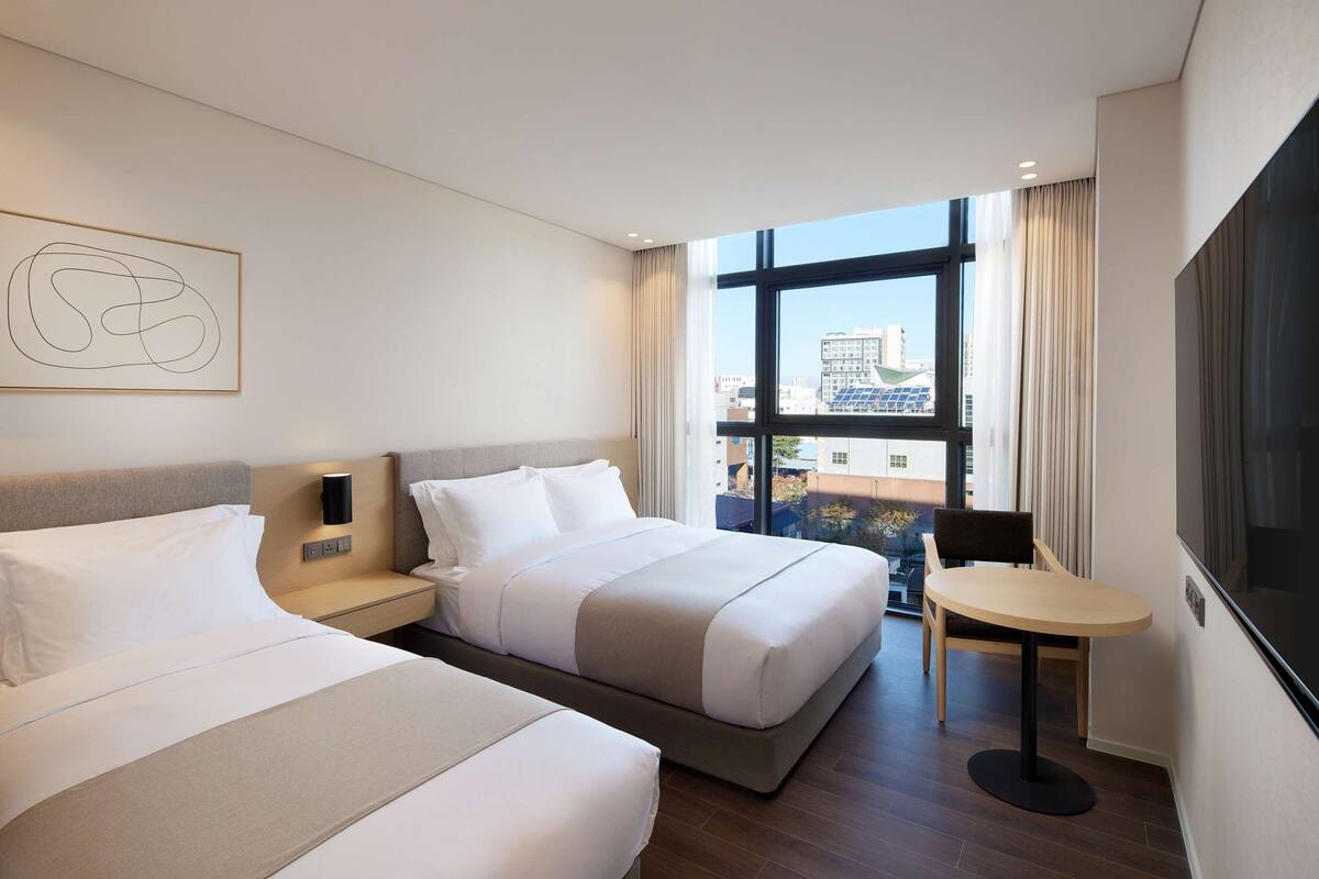 Quarto de hotel com duas camas, uma mesinha, uma televisão e janela com vista para a cidade.