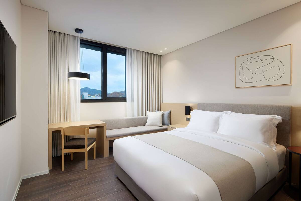 Quarto de hotel moderno com uma cama grande, uma escrivaninha e uma cadeira. Há uma janela com cortinas, uma obra de arte na parede e vista para as montanhas.