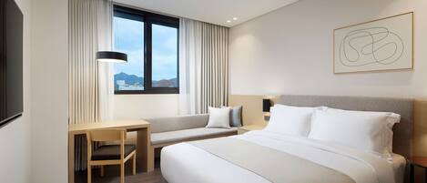 A modern hotel room with a large bed, a desk, and a chair. There is a window with curtains, a wall-mounted artwork, and a view of the mountains.