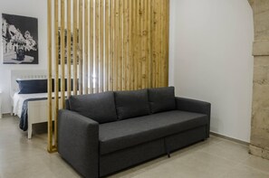 Un salon moderne avec un canapé gris, une cloison en lattes de bois, un lit avec une tête de lit blanche et une photographie en noir et blanc accrochée au mur.