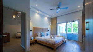 A modern bedroom with a large bed, a ceiling fan, and a view of the outdoors through a sliding glass door. - Modern 3 Bedroom Pool Villa - MR54 (Hua Hin)