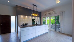 A modern kitchen with a central island, built-in appliances, and a dining area with a view of greenery. - Modern 3 Bedroom Pool Villa - MR54 (Hua Hin)