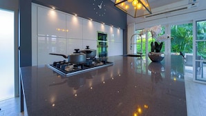 A modern kitchen with a central island, stainless steel appliances, and a large window with a view of greenery. - Modern 3 Bedroom Pool Villa - MR54 (Hua Hin)