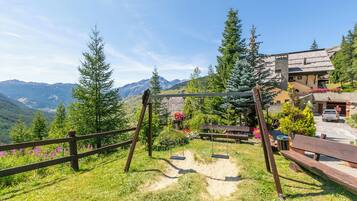 A wooden swing set, a bench, and a paved path in a garden with mountains in the background.