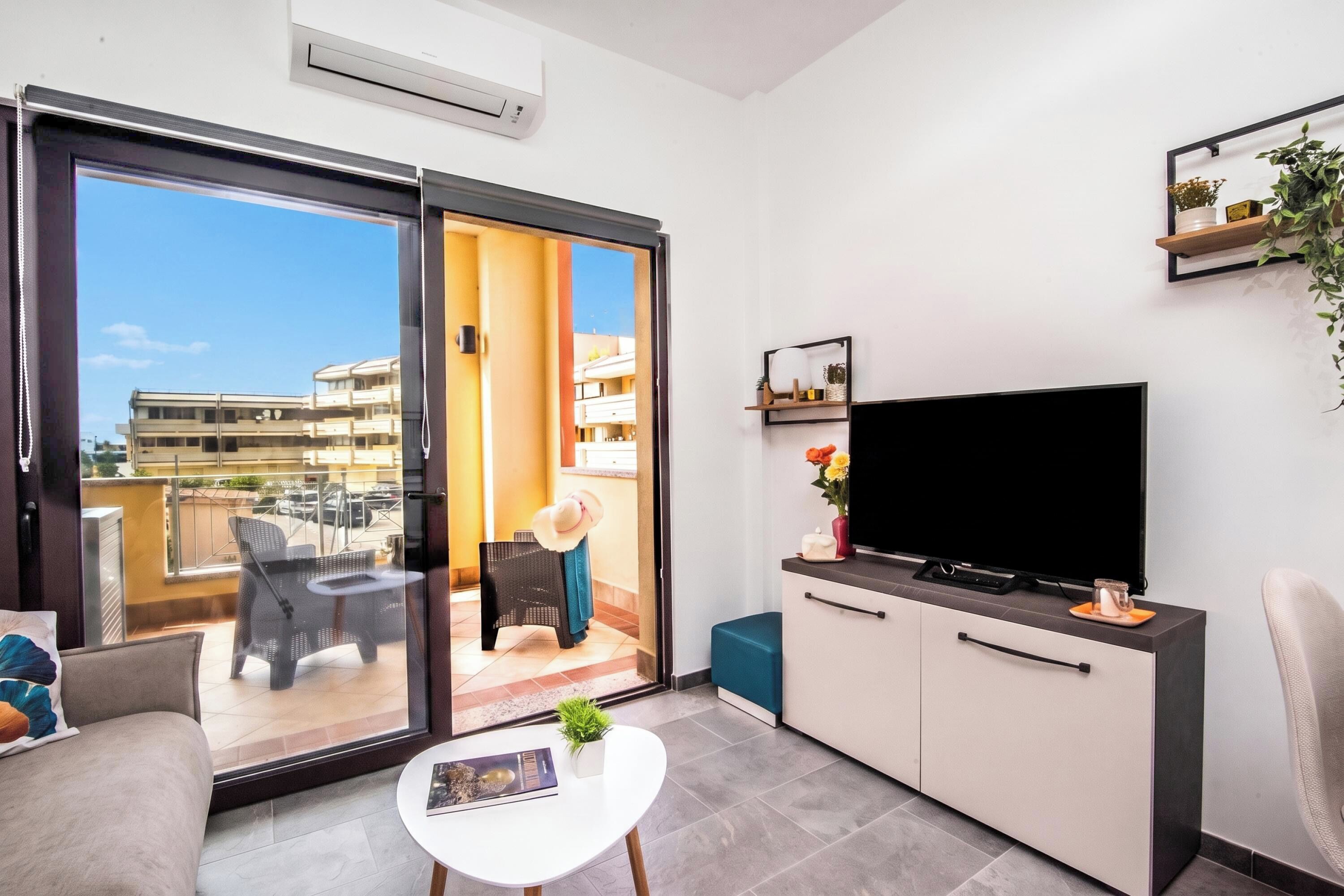 A modern living room with a sliding glass door to a balcony, a flat-screen TV, a small white table, and a sofa.