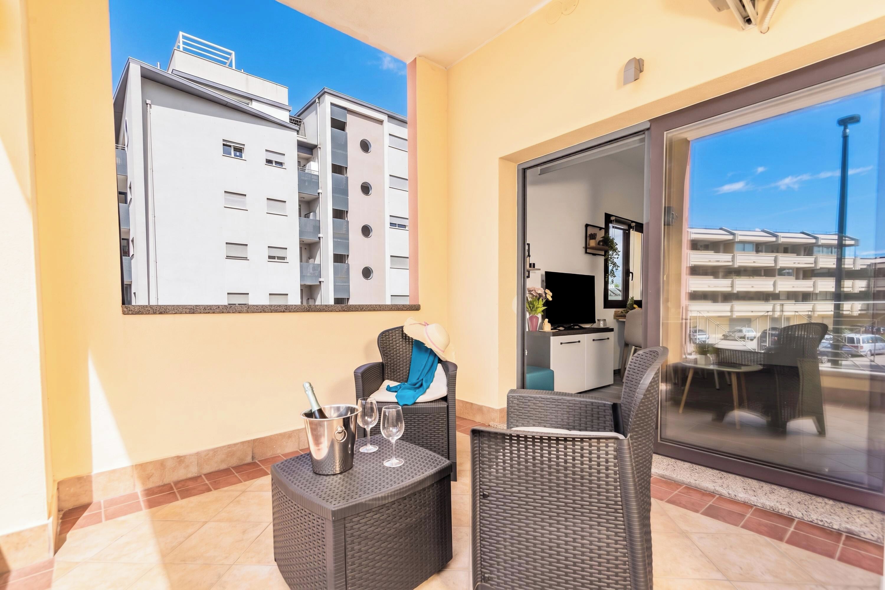 A balcony with wicker furniture, a champagne bottle, and glasses, overlooking a building with balconies.