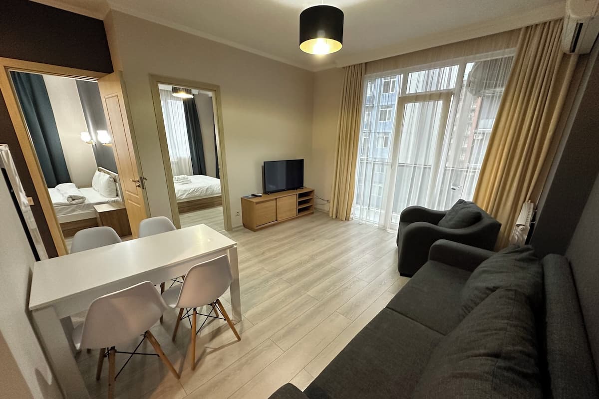 Ein modernes Hotelzimmer mit Essbereich, einer Couch, einem Fernseher und einem großen Fenster mit Vorhängen.