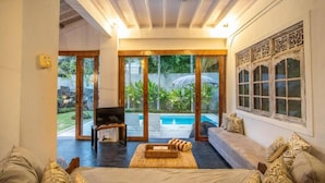 A spacious living area with a large window, a sofa, a TV, and a small table. - Villa Dolcevita Seminyak  (Seminyak)