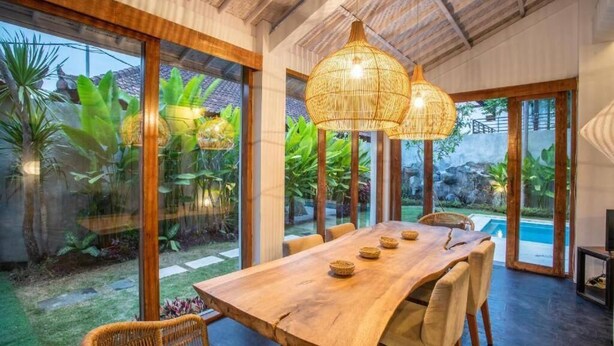 A spacious dining area with a large wooden table and wicker chairs, surrounded by large windows offering views of a lush garden and a swimming pool. - Villa Dolcevita Seminyak  (Seminyak)