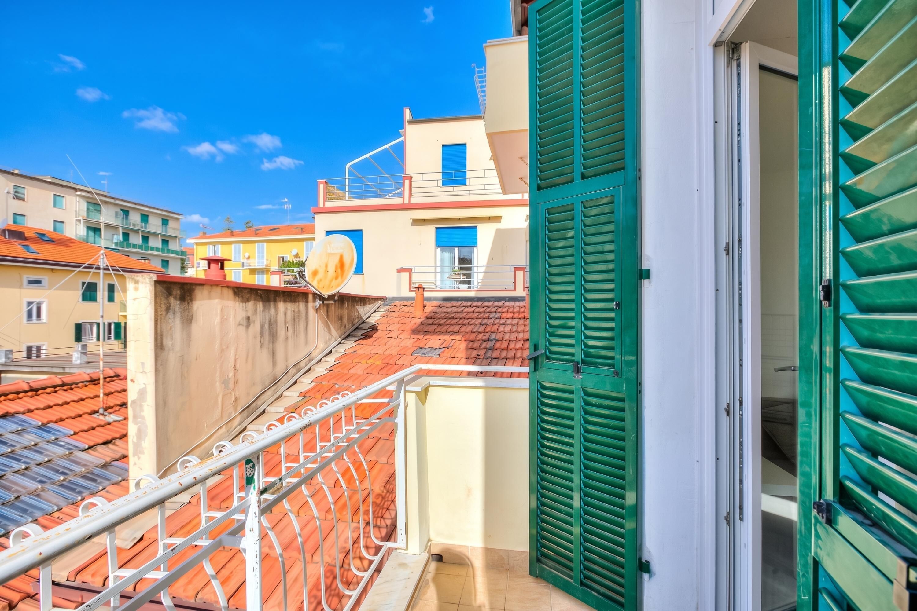 A balcony with a metal railing, a door with green shutters, and a view of buildings with red roofs.