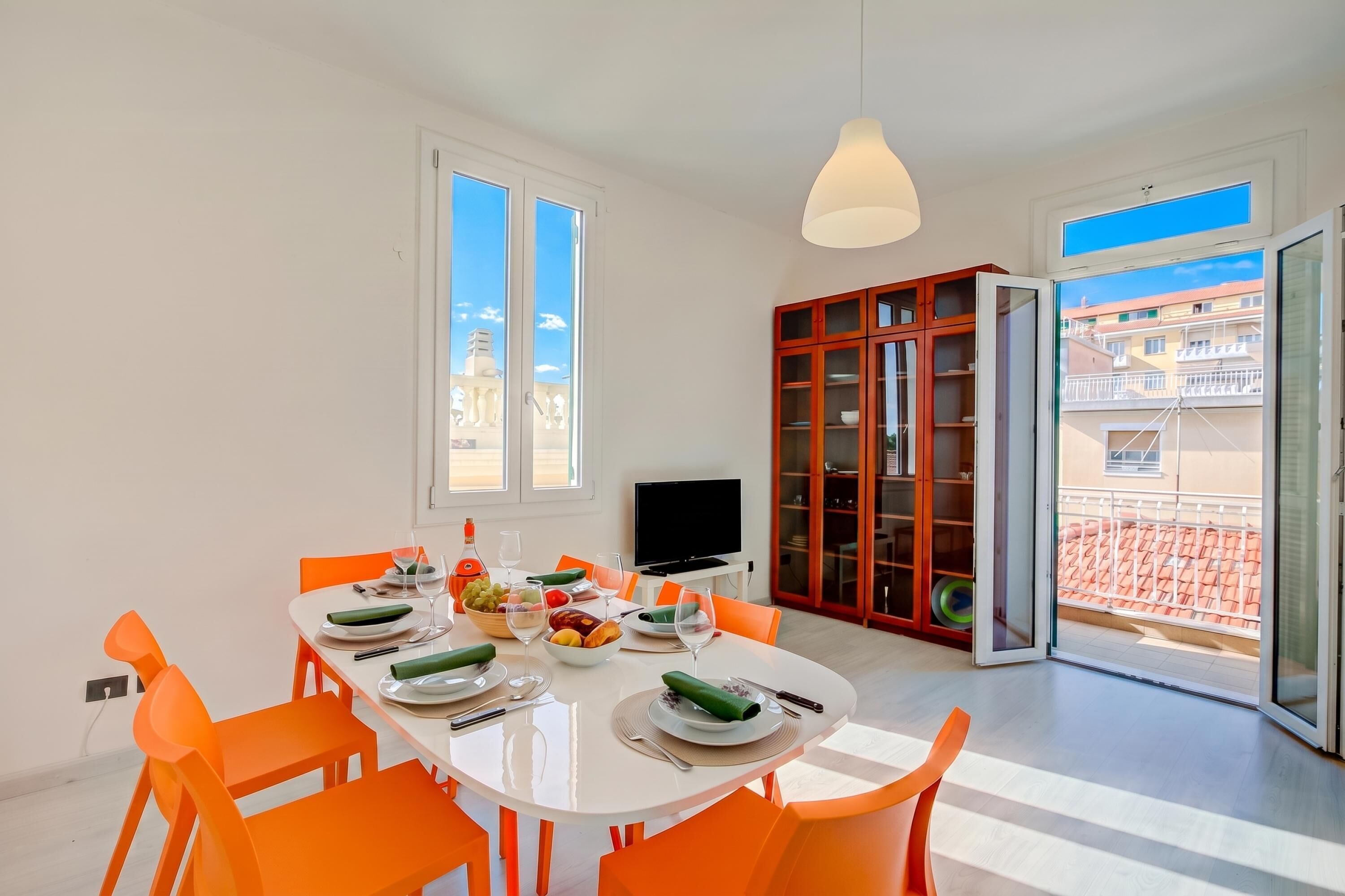 Comedor con una mesa blanca, sillas naranjas y vista a un edificio a través de la puerta abierta.