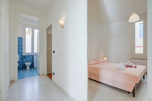 A bedroom with a bed, a bedside table, a window, and a bathroom with a toilet and sink.