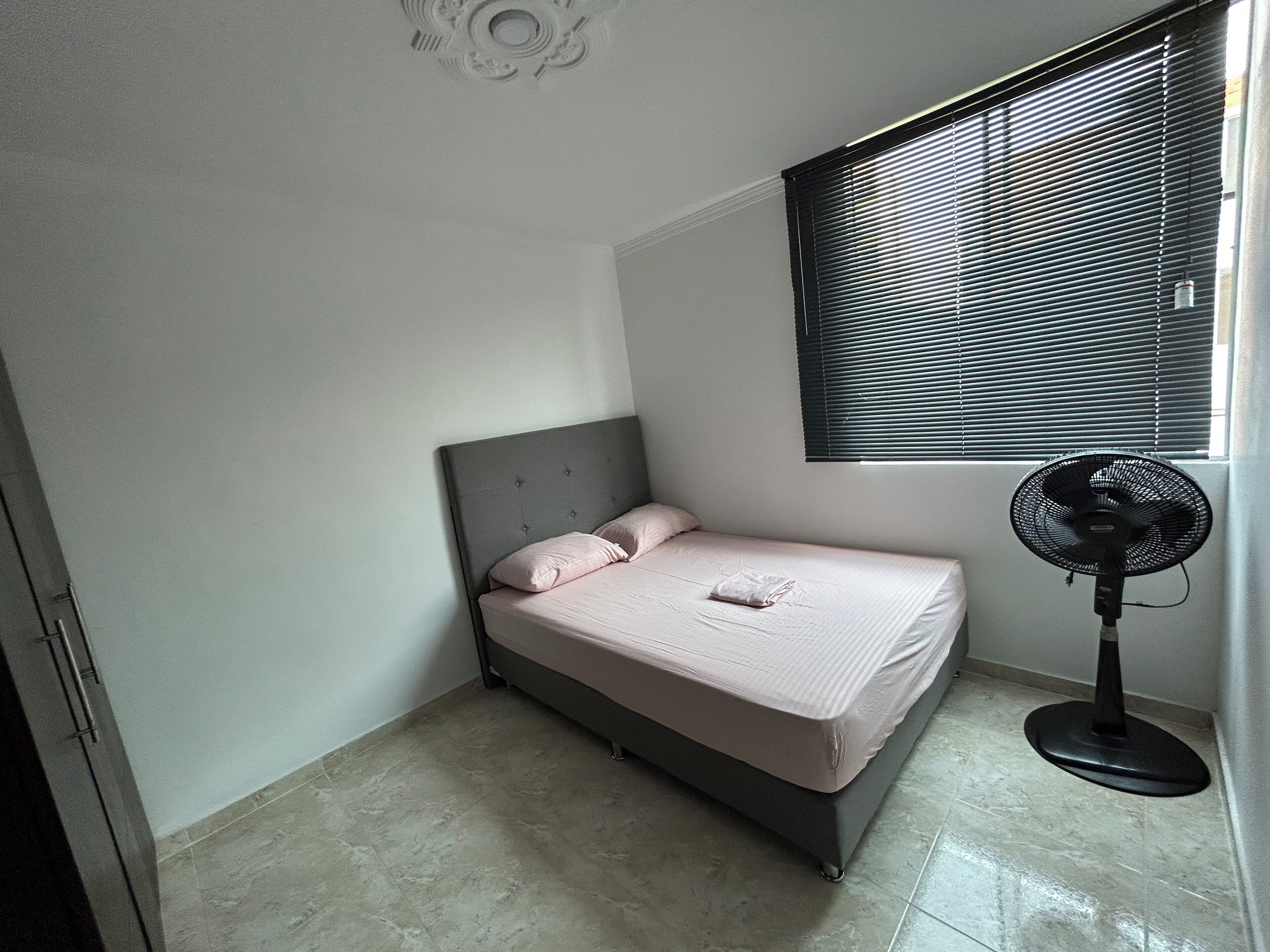 Un dormitorio pequeño y ordenado con cama, ventana con persianas y ventilador de pie.