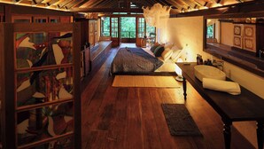 A spacious room with a high ceiling, wooden beams, a large bed, a bedside table, a window with a view, and a wall with artwork. - Sama Retreats - Kosgoda (Kosgoda)