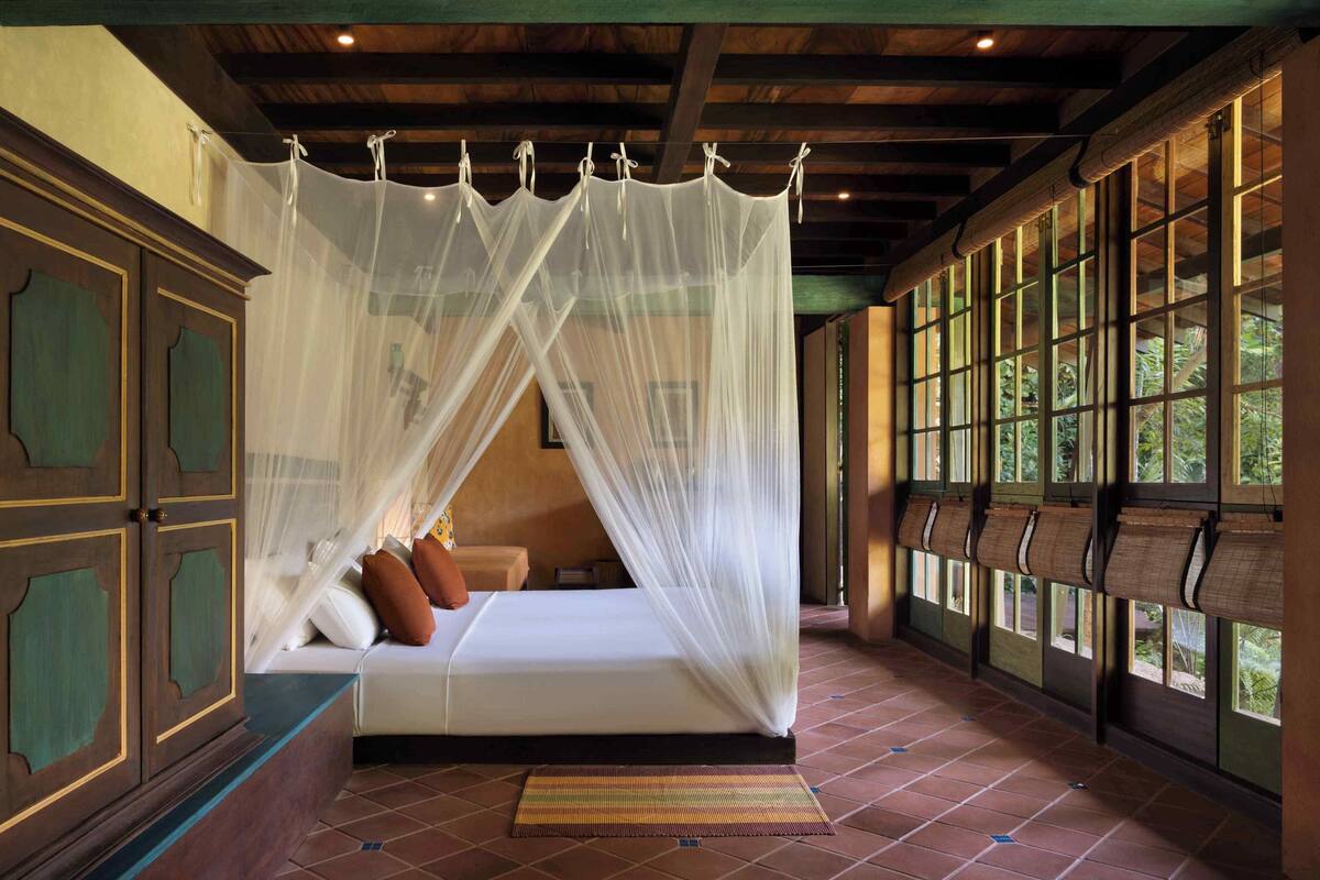 Une chambre avec un lit sous un baldaquin blanc, des meubles en bois et de grandes fenêtres équipées de stores.