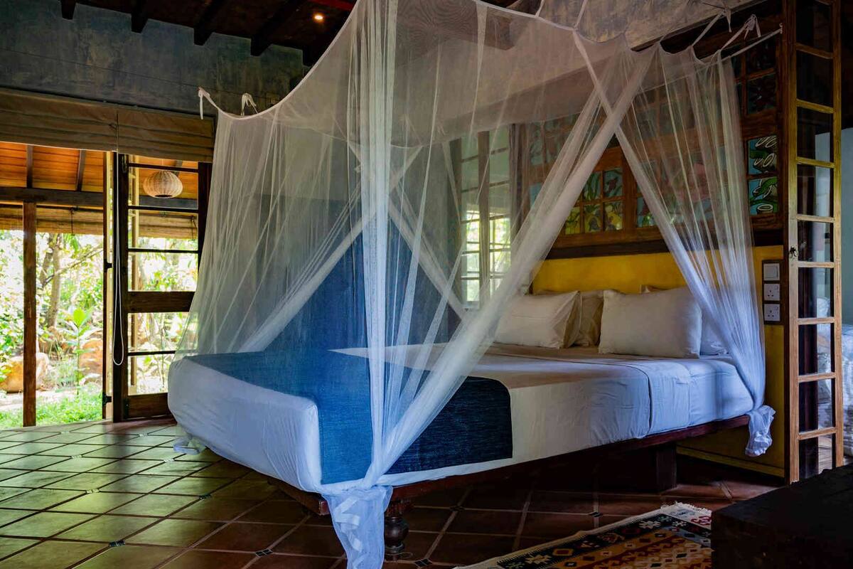 Une chambre avec un lit sous un dais, de grandes fenêtres et une vue sur la verdure environnante.