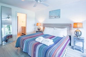 A bedroom with a large bed, two bedside lamps, a ceiling fan, a window with blinds, a door leading to another room, and a painting on the wall.