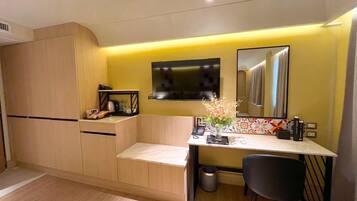 A compact hotel room with a kitchenette, a flat-screen TV mounted above a mirror, and a small desk with a chair.