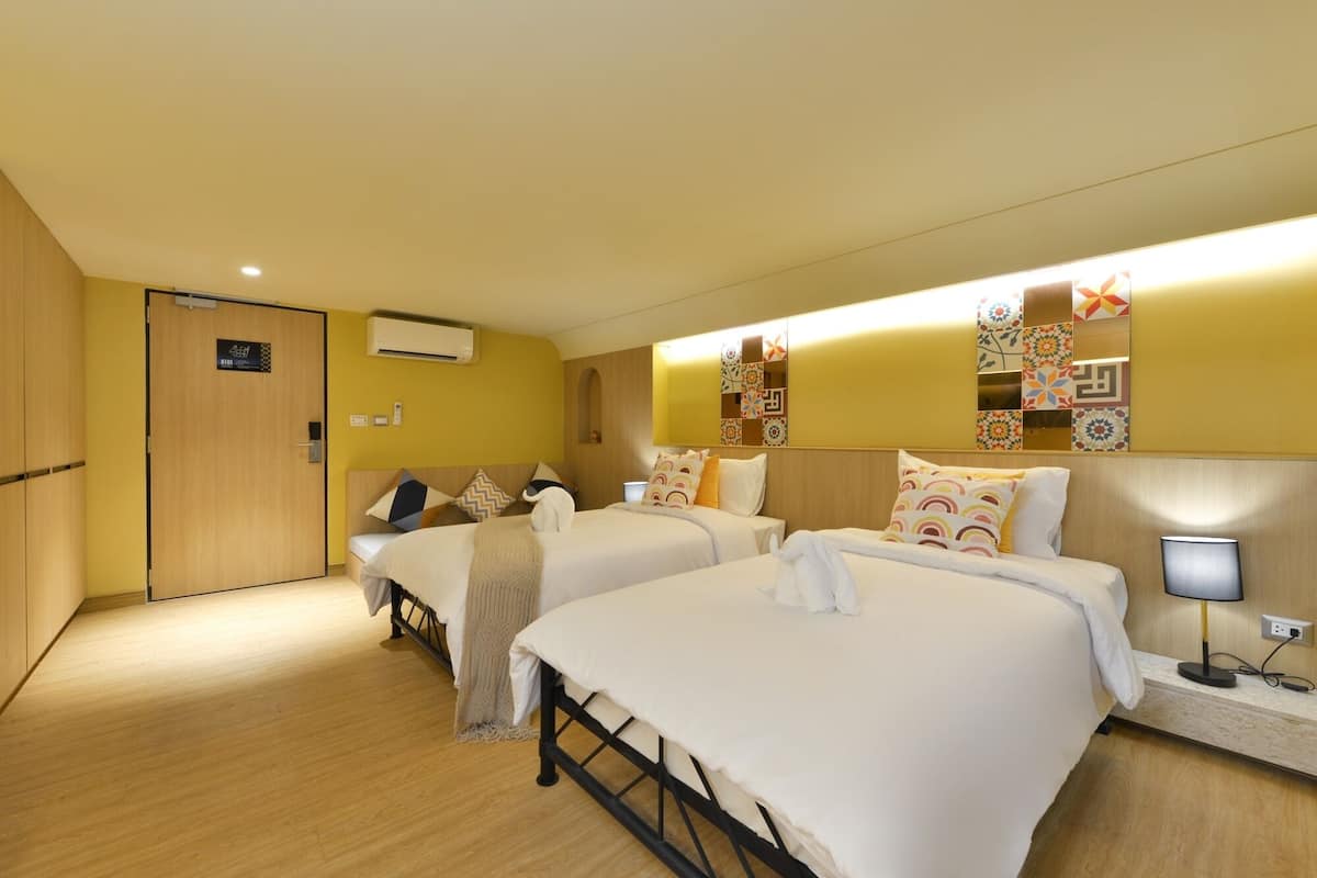Une chambre d’hôtel avec deux lits, un plancher en bois, un climatiseur fixé au mur et une table de chevet avec une lampe.