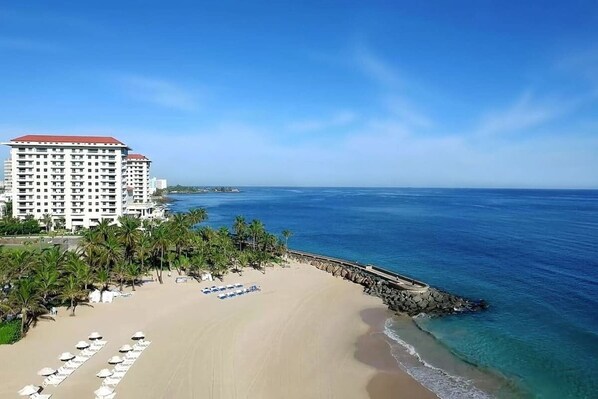 A beach with a hotel, palm trees, and a clear blue sky.