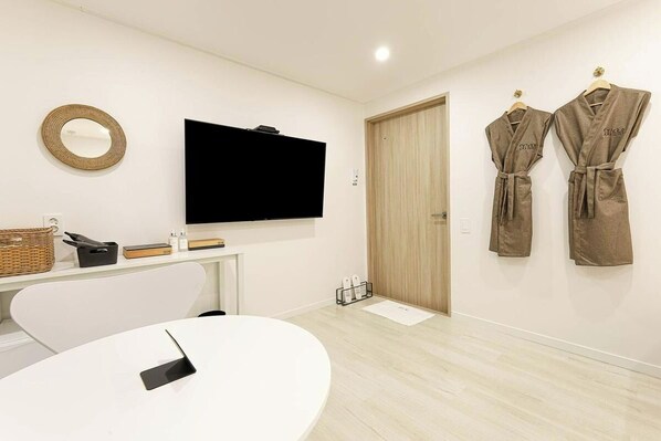 A modern room with a flat-screen TV, a round mirror, a white table with a smartphone, and two robes hanging on the wall. - hotel daor (Busan)
