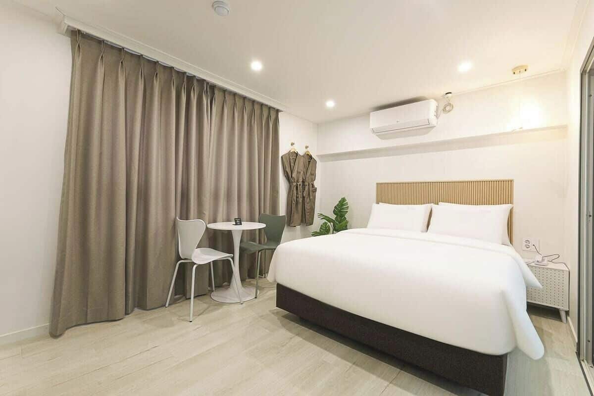A hotel room with a large bed, a small table with chairs, a plant, and an air conditioning unit.