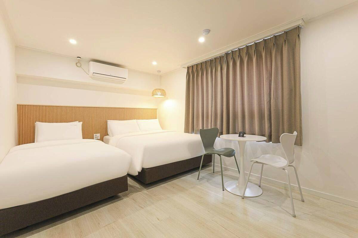 A hotel room with two beds, a small table with chairs, an air conditioning unit, and a window with curtains.