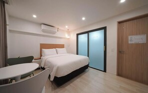 A hotel room with a bed, a small table, a chair, a wardrobe, and an air conditioning unit. - hotel daor (Busan)