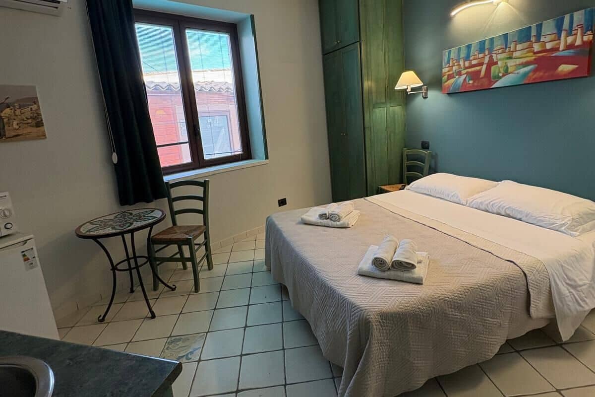 Ein kleines, sauberes Zimmer mit einem Bett, einem Tisch mit Stuhl, einem Fenster und einem Bild an der Wand.
