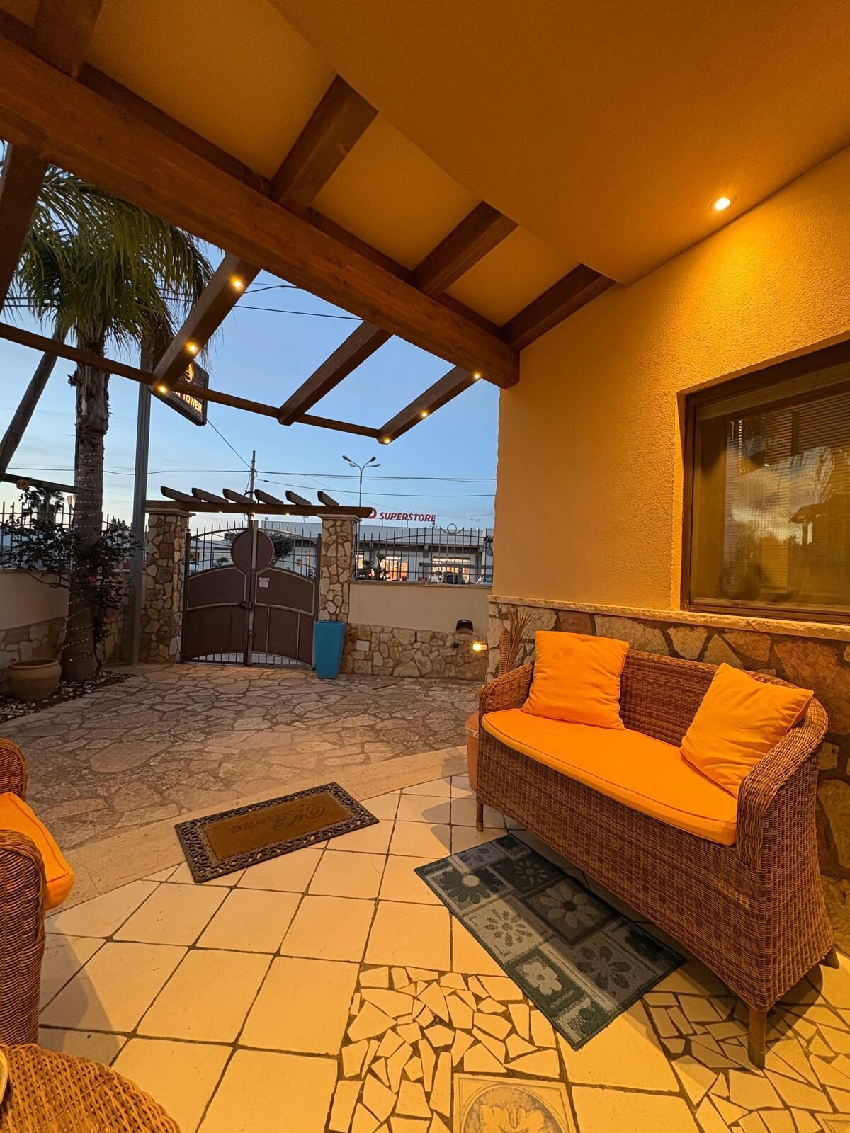 A covered outdoor patio with a wicker sofa, a stone wall, and a Superstore entrance in the background.