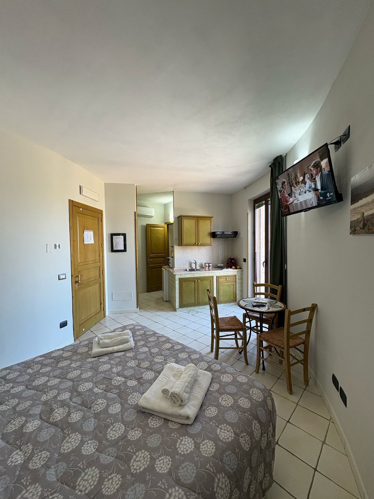 A bedroom with a bed, a small dining table with chairs, a kitchen area, and a TV mounted on the wall.