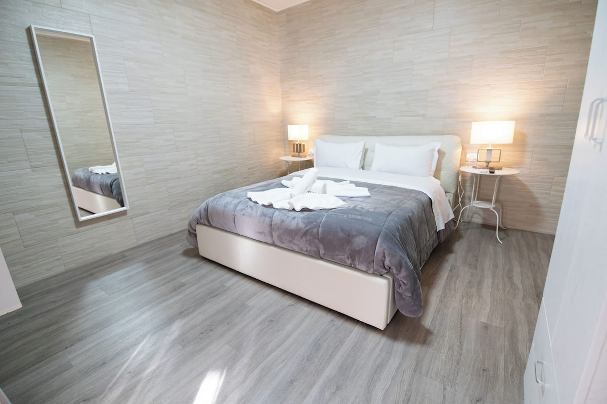 Moderni hotellihuone, jossa on suuri sänky, kaksi yöpöytää valaisimineen, suuri peili ja puulattia.