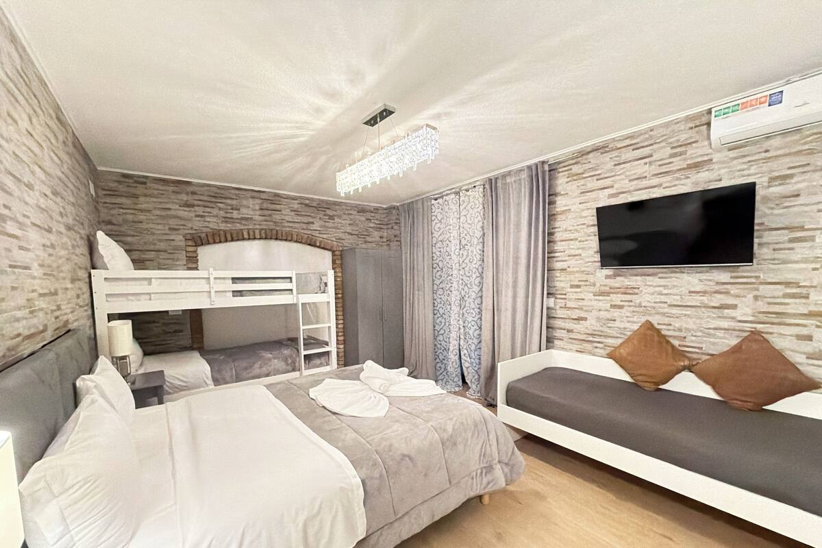 Hotellihuone, jossa on kerrossänky, yksi erillinen sänky, taulutelevisio ja kivipintainen seinä.