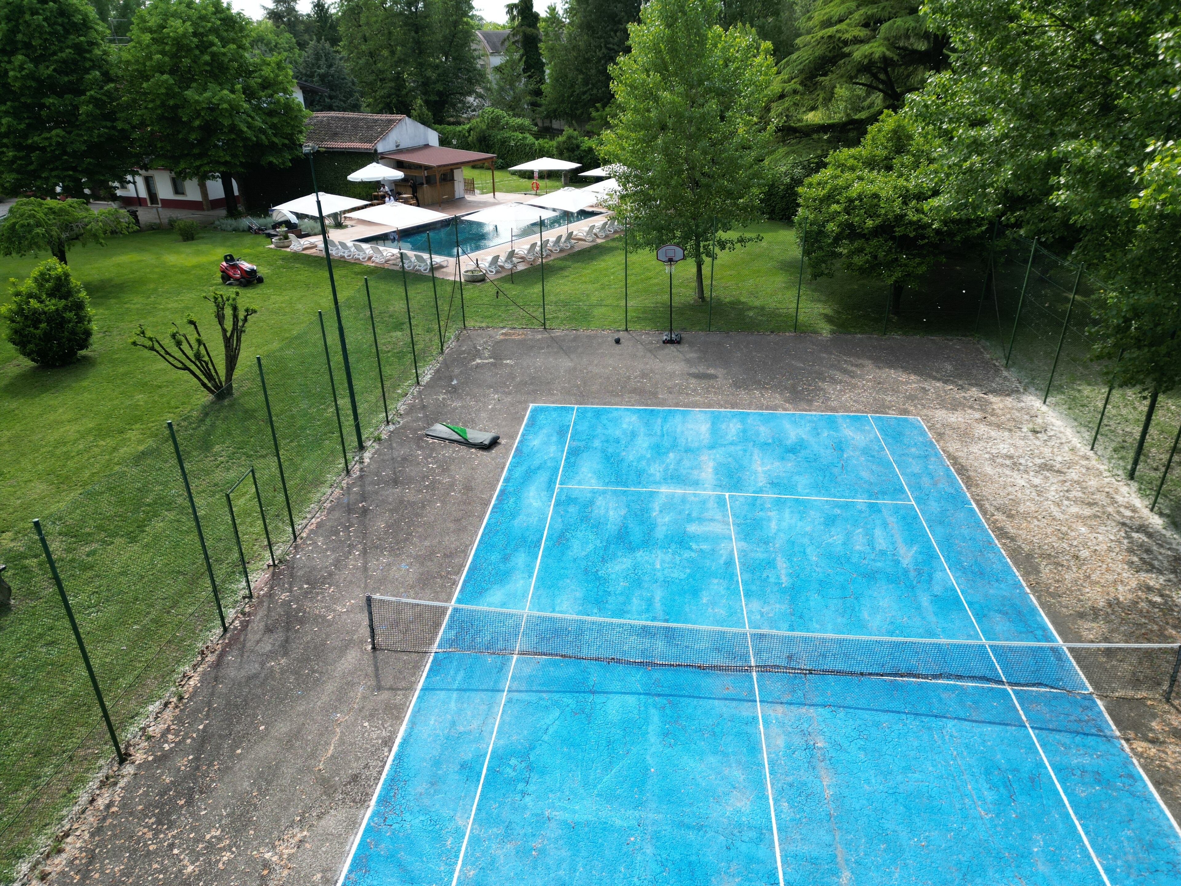 Ein blauer Tennisplatz mit Netz, umgeben von einem Zaun, und im Hintergrund ein kleiner Poolbereich.
