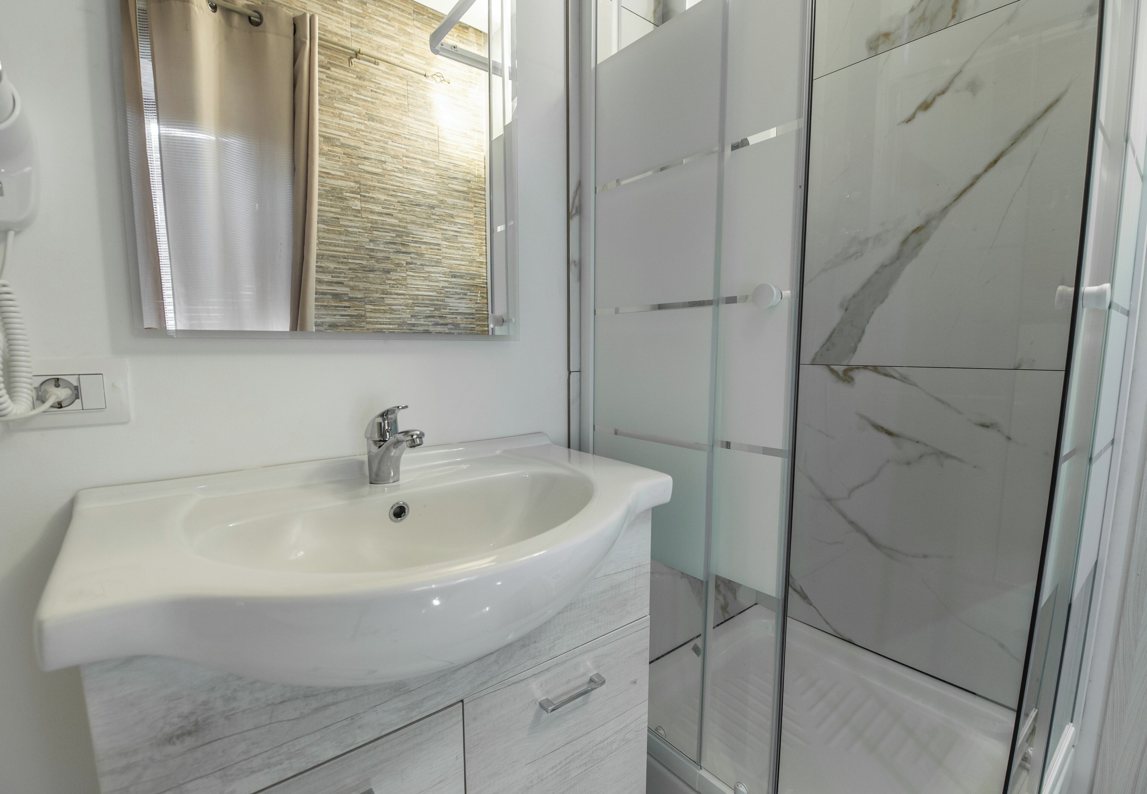 Un baño con lavamanos blanco, un espejo, ducha con puertas de vidrio y pared con textura de madera.