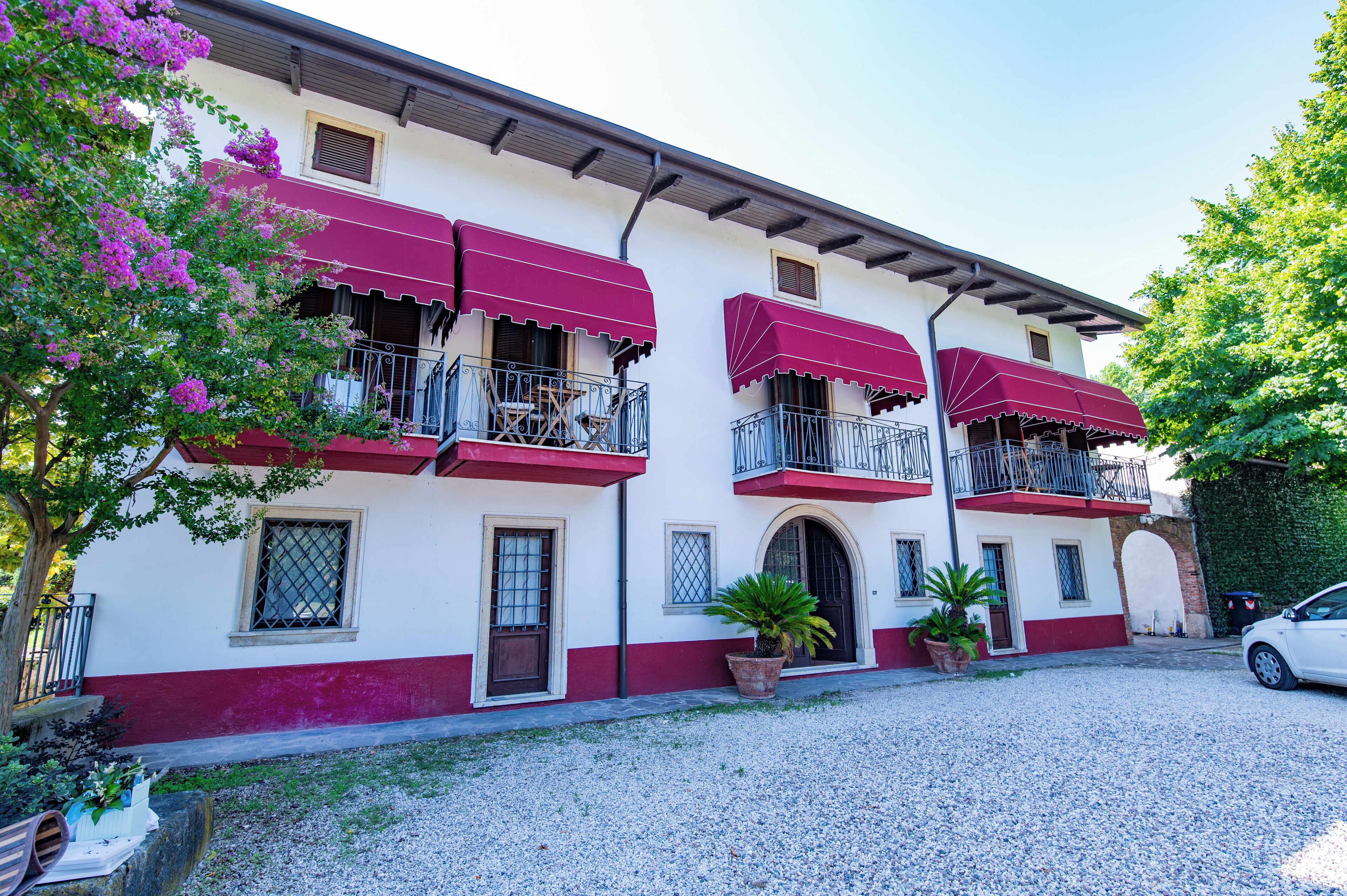 A building with a white facade and red accents, featuring balconies with red awnings, potted plants, and a gravel driveway.