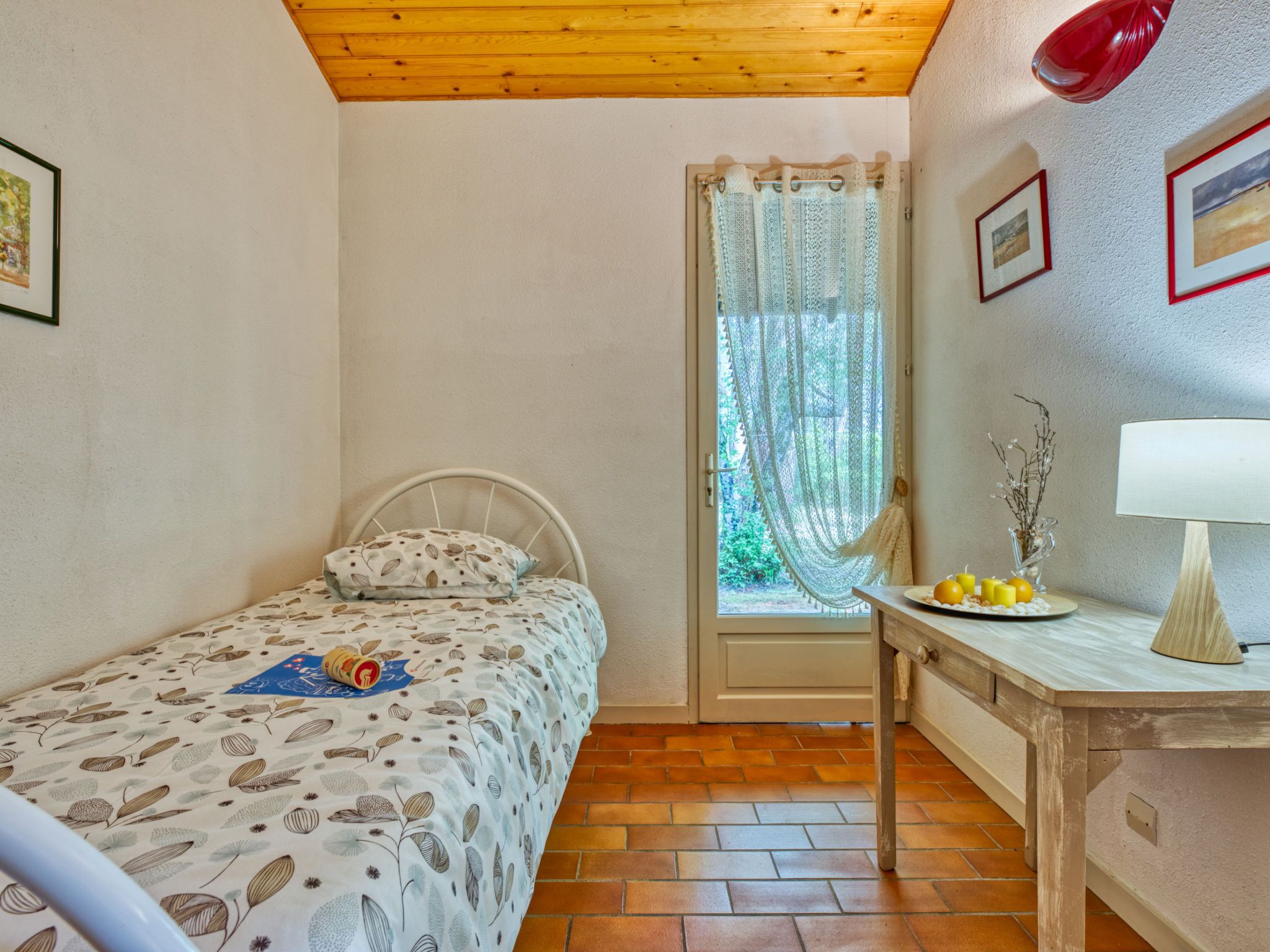 Une petite chambre avec un lit simple, un plafond en bois, une fenêtre avec un rideau, une table avec une lampe et un mur orné de tableaux.