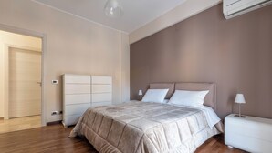 Ένα σύγχρονο υπνοδωμάτιο με ξύλινο δάπεδο, ένα κρεβάτι με μπεζ κουβέρτα, ένα λευκό κομοδίνο και μια μονάδα κλιματισμού στον τοίχο.