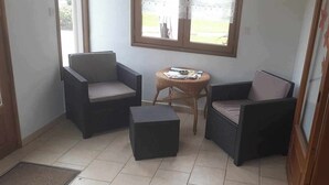 A corner room with two wicker chairs, a small round table, and a window with a view of the outdoors. - Gîte Chez Mamie 
A pleasant and quiet cottage by the sea (Saint-Marcouf)