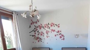 A bedroom with two beds, a wooden dresser, a window with curtains, a floral wall decoration, and a ceiling light fixture. - Gîte Chez Mamie 
A pleasant and quiet cottage by the sea (Saint-Marcouf)