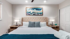 A bedroom with a large bed, two bedside tables, a ceiling fan, and a painting on the wall.